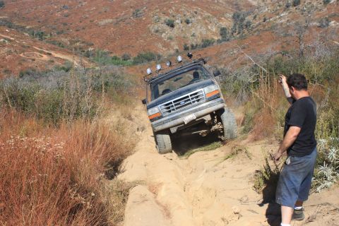 1992 Ford Bronco - A 1992 Ford Bronco tackles a challenging off-road trail, its tires digging into the sandy terrain as a bystander observes.
