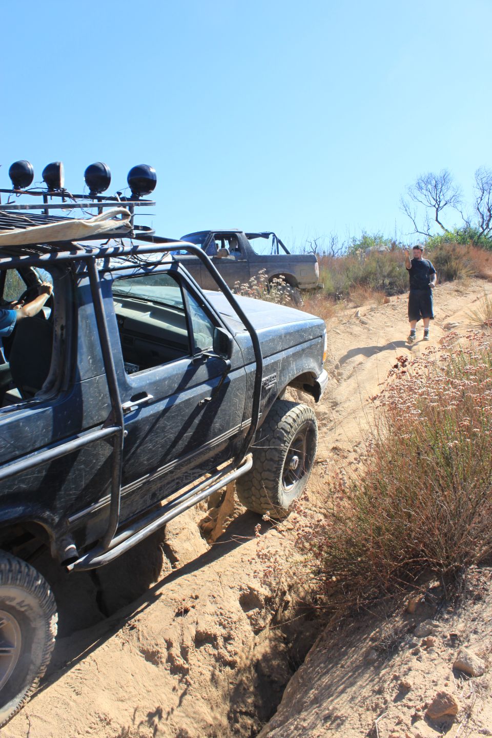 Two off-road vehicles navigate a steep, sandy trail with brush and trees on the sides. A person walks up the trail behind the lead vehicle