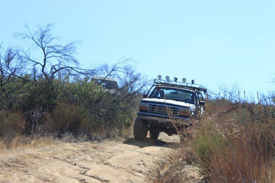 White 1992 Ford Bronco with roof lights driving on a dirt trail, another vehicle visible behind