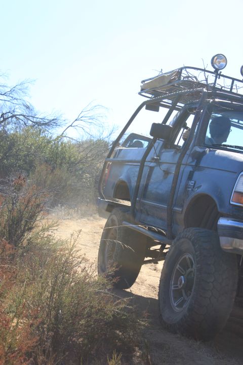 1992 Ford Bronco - Modified 1992 Ford Bronco navigates a dusty off-road path with brush on either side.