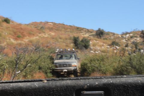 1992 Ford Bronco - A 1992 Ford Bronco navigates a rough, off-road path surrounded by dense vegetation and arid hills under a clear blue sky.