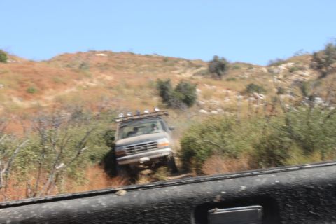 1992 Ford Bronco - A 1992 Ford Bronco navigates a , dry, hilly terrain, equipped with a roof rack and auxiliary lights.