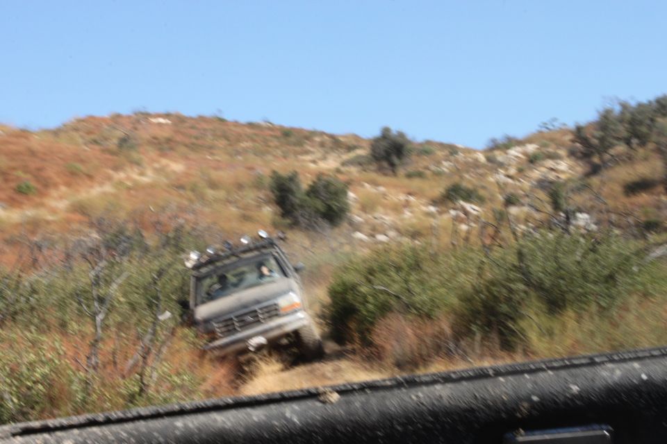 Off-road vehicle driving up a dirt hill with brush and trees. It has a roof rack with lights