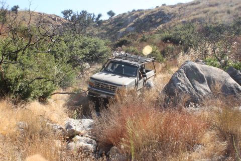 1992 Ford Bronco - A tan 1992 Ford Bronco navigates a challenging off-road trail surrounded by dry brush and large rocks.
