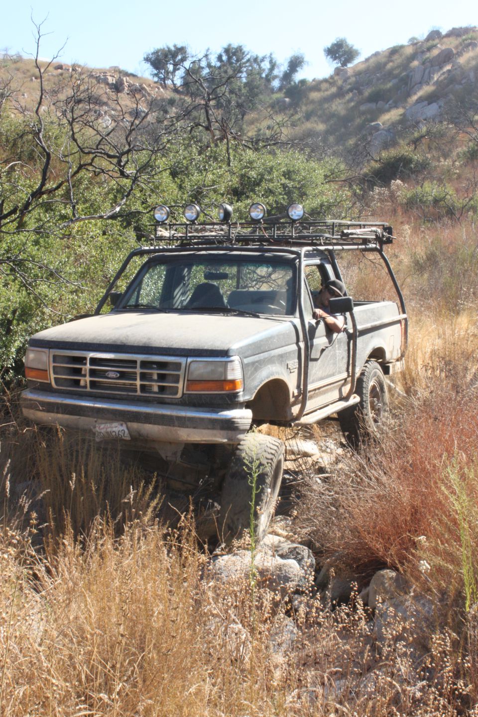 1992 Ford Bronco with roof rack and off-road lights driving over rocks and dry grass