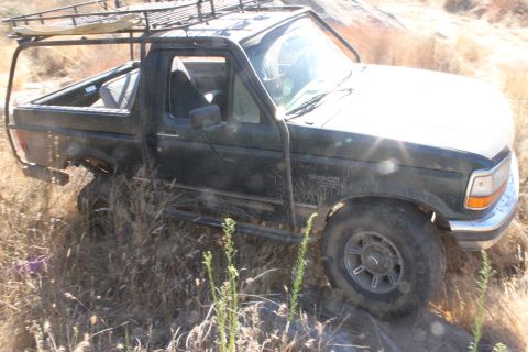 1992 Ford Bronco - A dark 1992 Ford Bronco is parked off-road, equipped with a roof rack and roll cage.