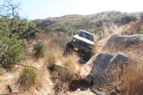 1992 Ford Bronco - A white SUV navigates a challenging rocky trail with dry brush and large boulders.