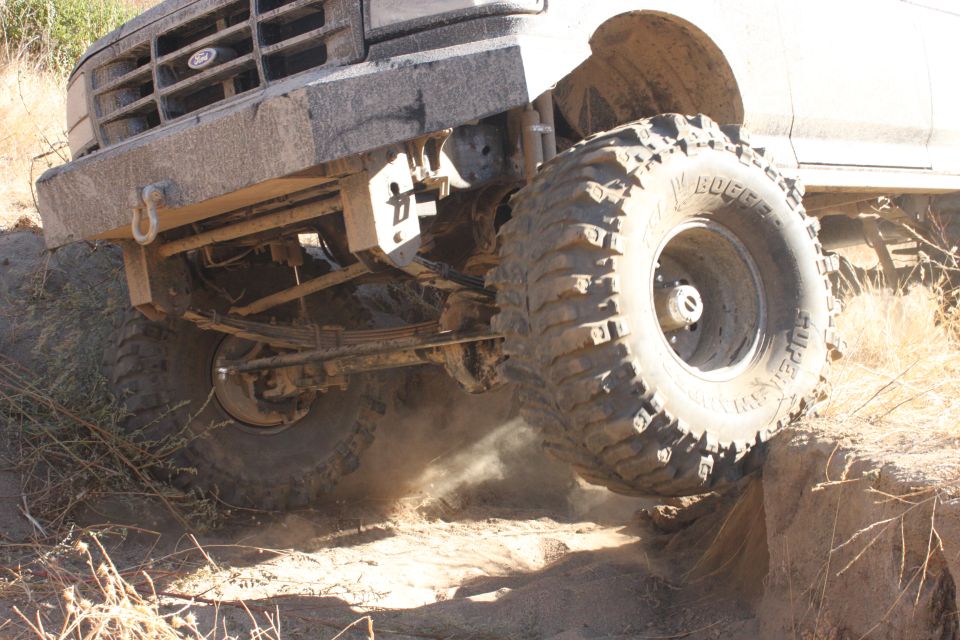 Mud-covered Ford truck front end with large off-road tires climbing over a rock