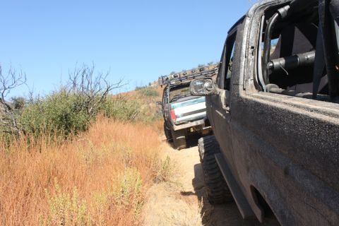 1992 Ford Bronco - Two off-road vehicles navigate a dusty trail through dry brush under a clear blue sky.