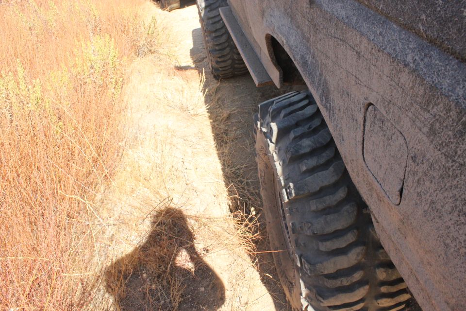 Off-road tire and fender of a vehicle, with dry brush and a shadow in the foreground. Supermotors.net logo visible