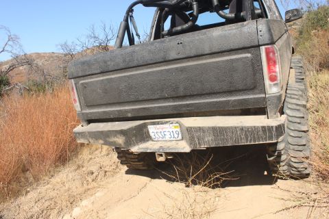 1992 Ford Bronco - Rear view of a black SUV with oversized tires, parked on a dusty trail under a clear blue sky.