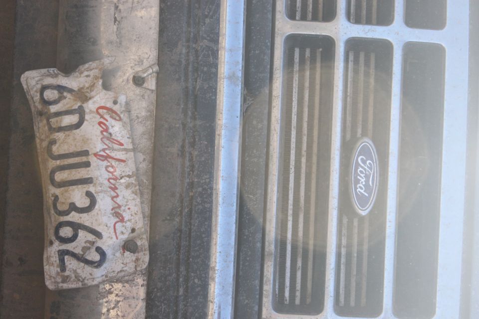 Close-up of a Ford emblem on a grille, with a dirty California license plate reading 6DJU362