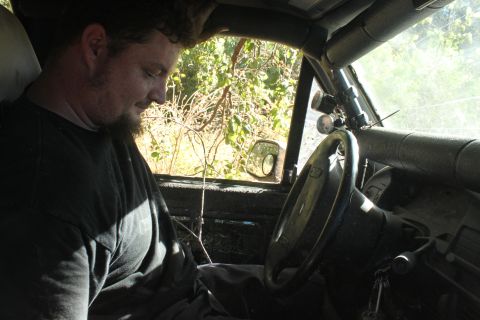 1992 Ford Bronco - Man sits inside a vehicle's interior, which features a roll cage and additional gauges, with foliage visible through the windows.