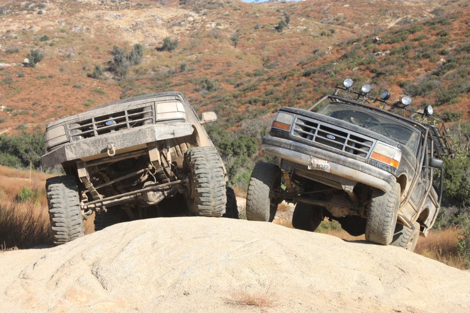 Two lifted Ford Broncos climbing a rocky hill off-road. One is tan, the other is black with extra lights