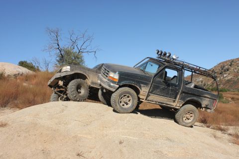 1992 Ford Bronco - Two off-road trucks, including a black 1992 Ford Bronco, navigate a steep, rocky incline under a clear blue sky.