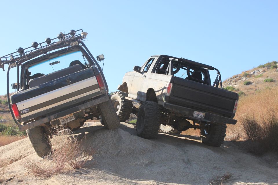 Two 1992 Ford Broncos climbing a rocky hill, one black and one tan. Both have off-road modifications