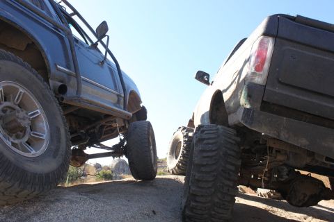 1992 Ford Bronco - Two heavily modified trucks are positioned on a rocky trail under a clear blue sky.
