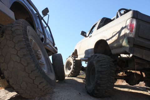 1992 Ford Bronco - Two heavily modified off-road trucks are parked side-by-side on a dirt trail under a clear blue sky.