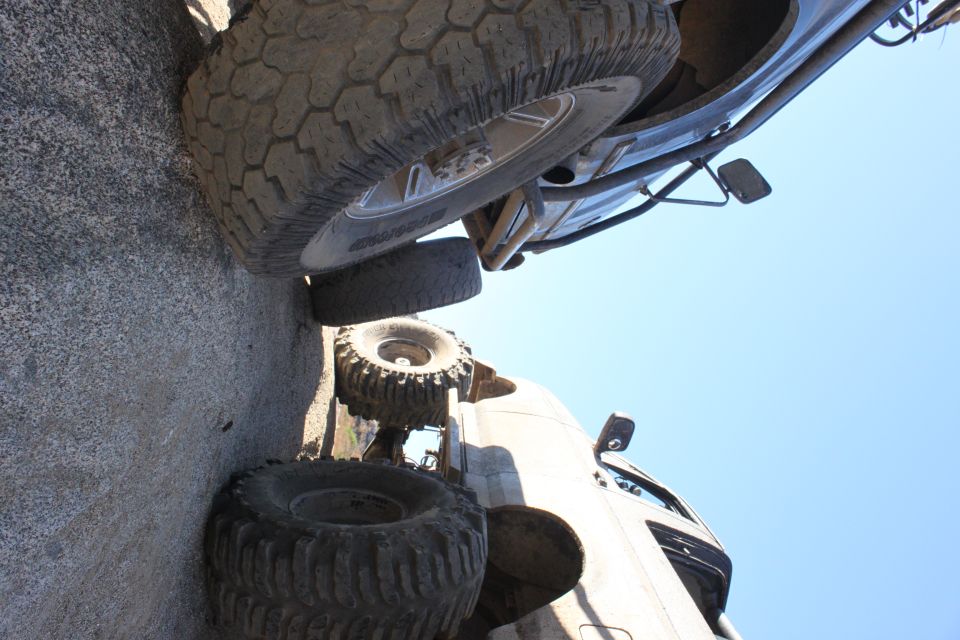 Two off-road vehicles with large tires on rocky ground under a clear blue sky. One vehicle is partially visible from underneath