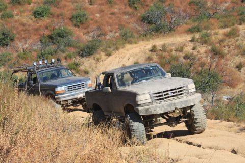1992 Ford Bronco - Two dusty Ford Broncos navigate a challenging off-road trail with dry brush and hills in the background.