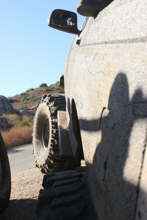 Mud-covered off-road vehicle side view, showing large tires and a side mirror. Sunny day with rocky hills in b