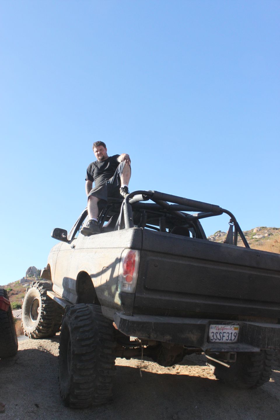 Lifted off-road truck with large tires, roll cage, and a man sitting on top. The vehicle is dusty and appears to be in a desert environment