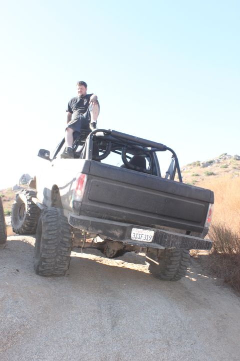 Rear view of a lifted, modified 1980s pickup truck with large tires and a roll cage. A man sits on the roof