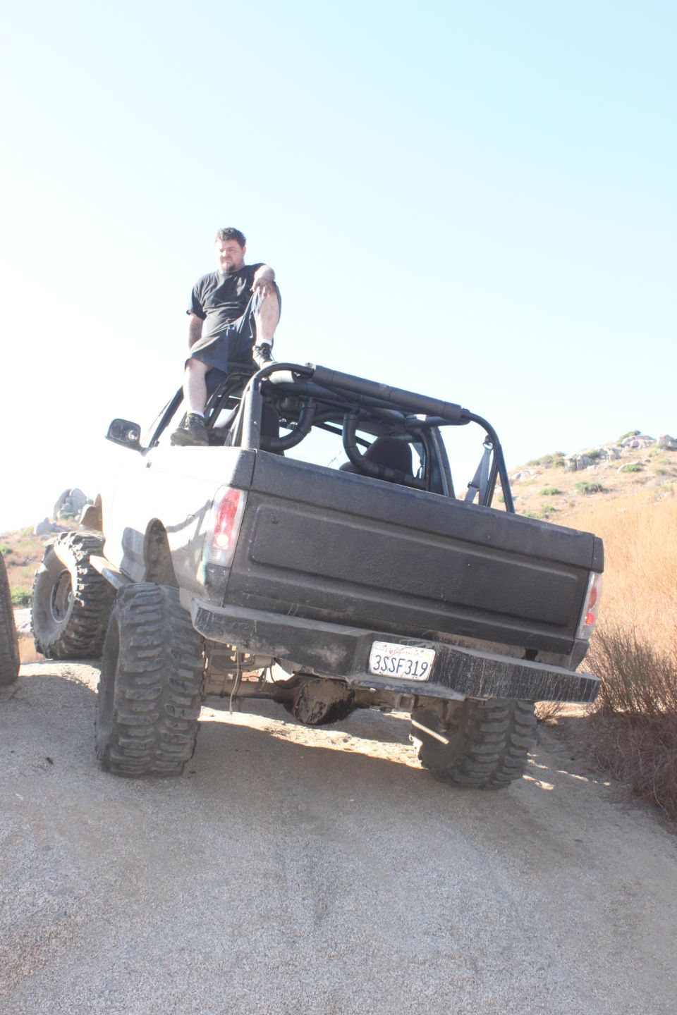 Rear view of a lifted, modified 1980s pickup truck with large tires and a roll cage. A man sits on the roof