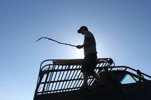 Person stands on roof rack of vehicle, holding a stick against a bright blue sky