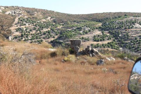 Side mirror reflection shows a dry, rocky hillside with sparse green trees and brown grass under a clear blue