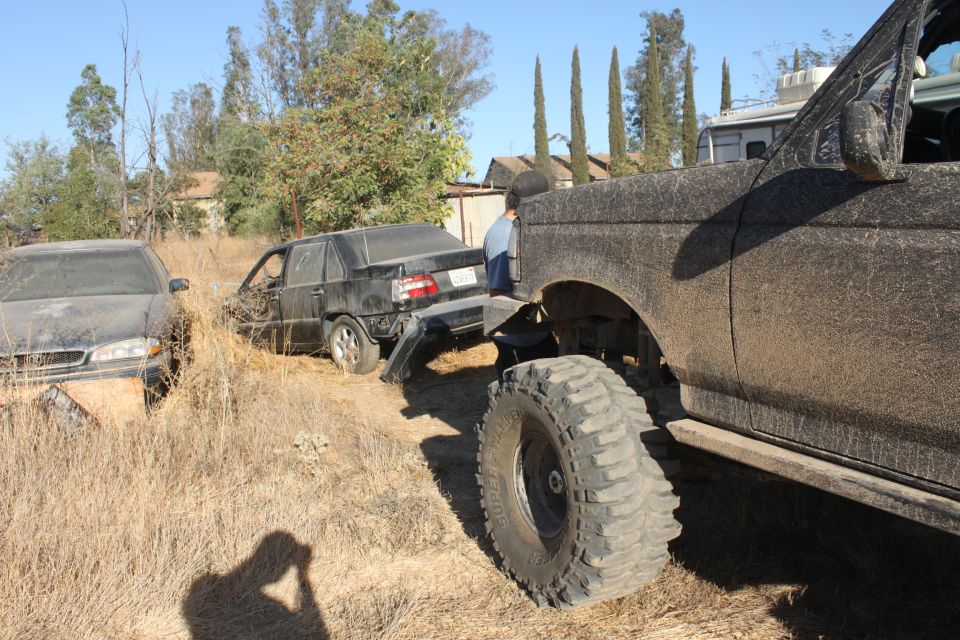 Muddy off-road truck pulling a wrecked black car out of dry grass. A person is visible in the truck's cab