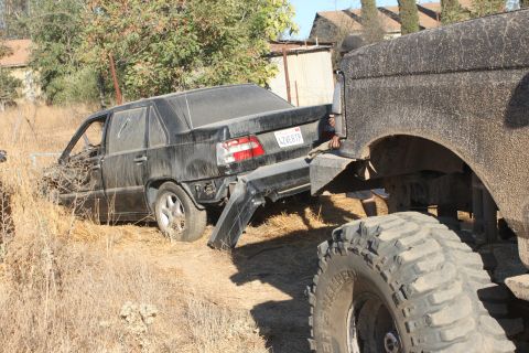 Black sedan's rear bumper is detached as it's towed by a large truck