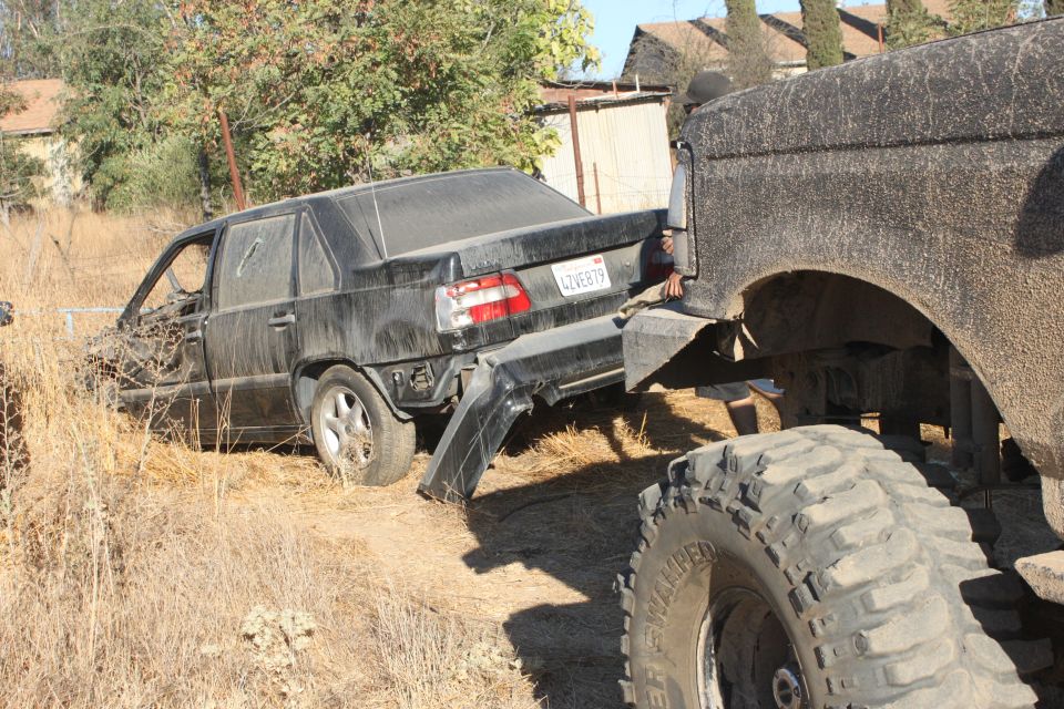Black sedan's rear bumper is detached as it's towed by a large truck