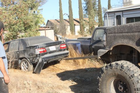 Wrecked black sedan with damaged bumper, being towed by a large truck with off-road tires. RV in background