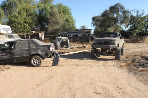 Ford F-150 being used to tow a damaged sedan in a junkyard. Another truck cab is visible in the background