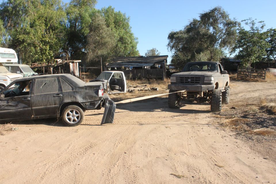 Lifted truck towing a wrecked sedan with a tow strap. Other vehicles and structures in background