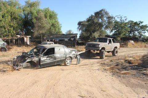 Lifted pickup truck towing a heavily damaged car on a dirt road near some old buildings