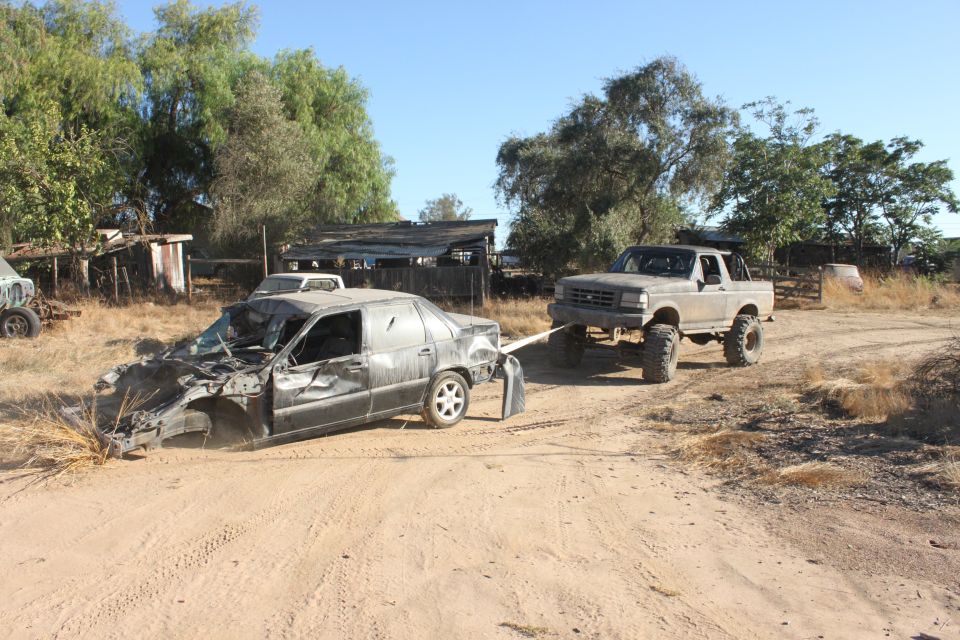 Gray 1992 Ford Bronco towing a heavily damaged black sedan on a dirt road