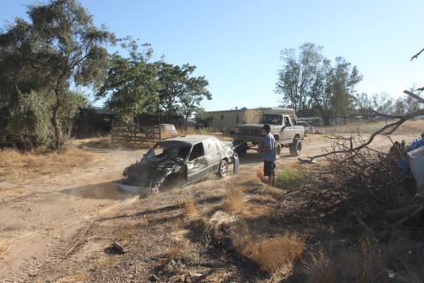 Wrecked sedan being towed by a lifted pickup truck on a dirt road. Person stands between vehicles
