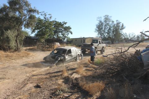 Guy with a lifted truck is towing a wrecked car on a dirt road. Some buildings and trees in the background