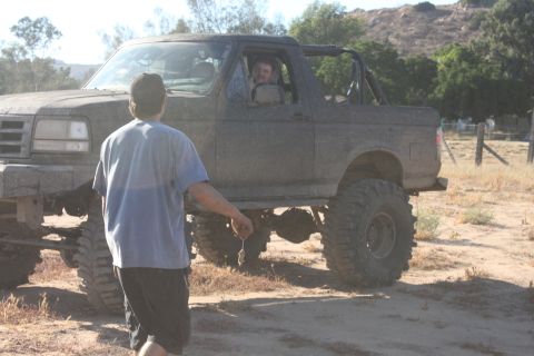 Muddy lifted SUV with large tires, person standing in front, another person in driver's seat