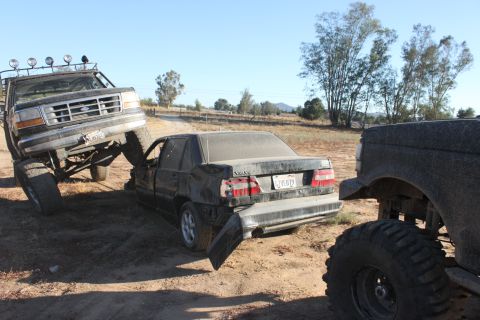 Big lifted truck on top of a wrecked sedan, another truck to the right
