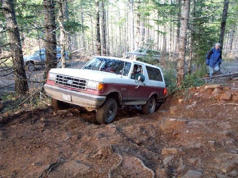 1982 Ford Bronco - A red and white Ford Bronco drives off-road through a wooded area with another vehicle in the background.