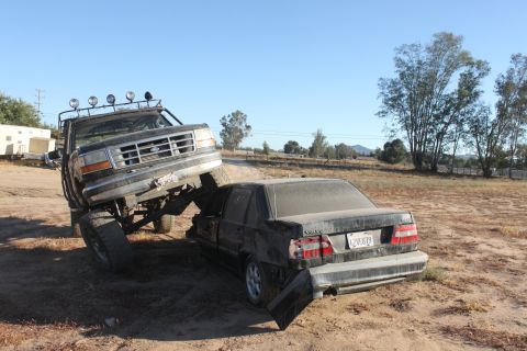 Lifted Ford truck on top of a dark sedan, crushing it. Truck has lights on roof. Sedan has license plate visib