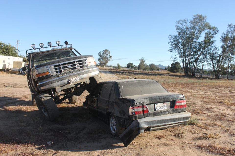 Lifted Ford truck on top of a dark sedan, crushing it. Truck has lights on roof. Sedan has license plate visible