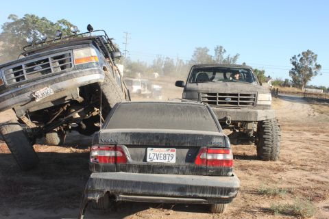 Two lifted Ford trucks are off-roading, one is climbing over a Volvo sedan
