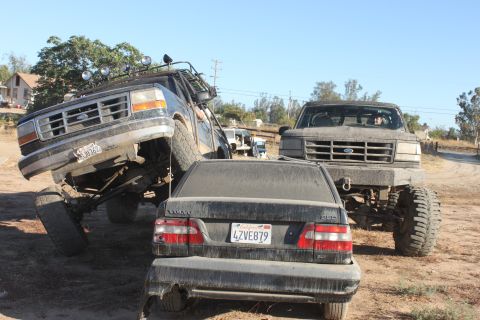 Two lifted Ford trucks, one with a roof rack, are parked around a dusty Volvo sedan. The truck on the left has