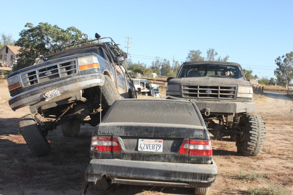 1992 Ford Bronco lifted with off-road lights, front grille visible, next to another lifted Ford truck and a black Volvo sedan