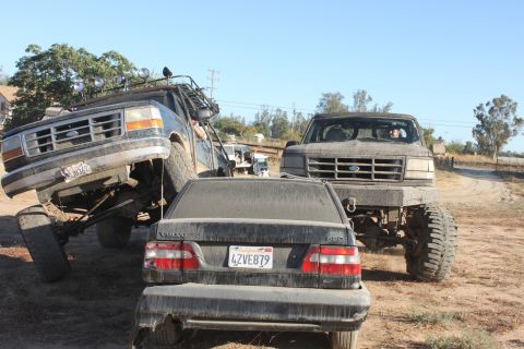 two lifted Ford trucks, one on its side, next to a Volvo sedan on a dirt road