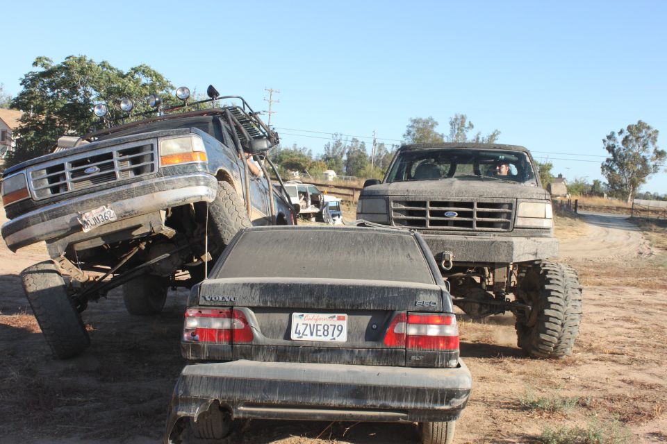 two lifted Ford trucks, one on its side, next to a Volvo sedan on a dirt road